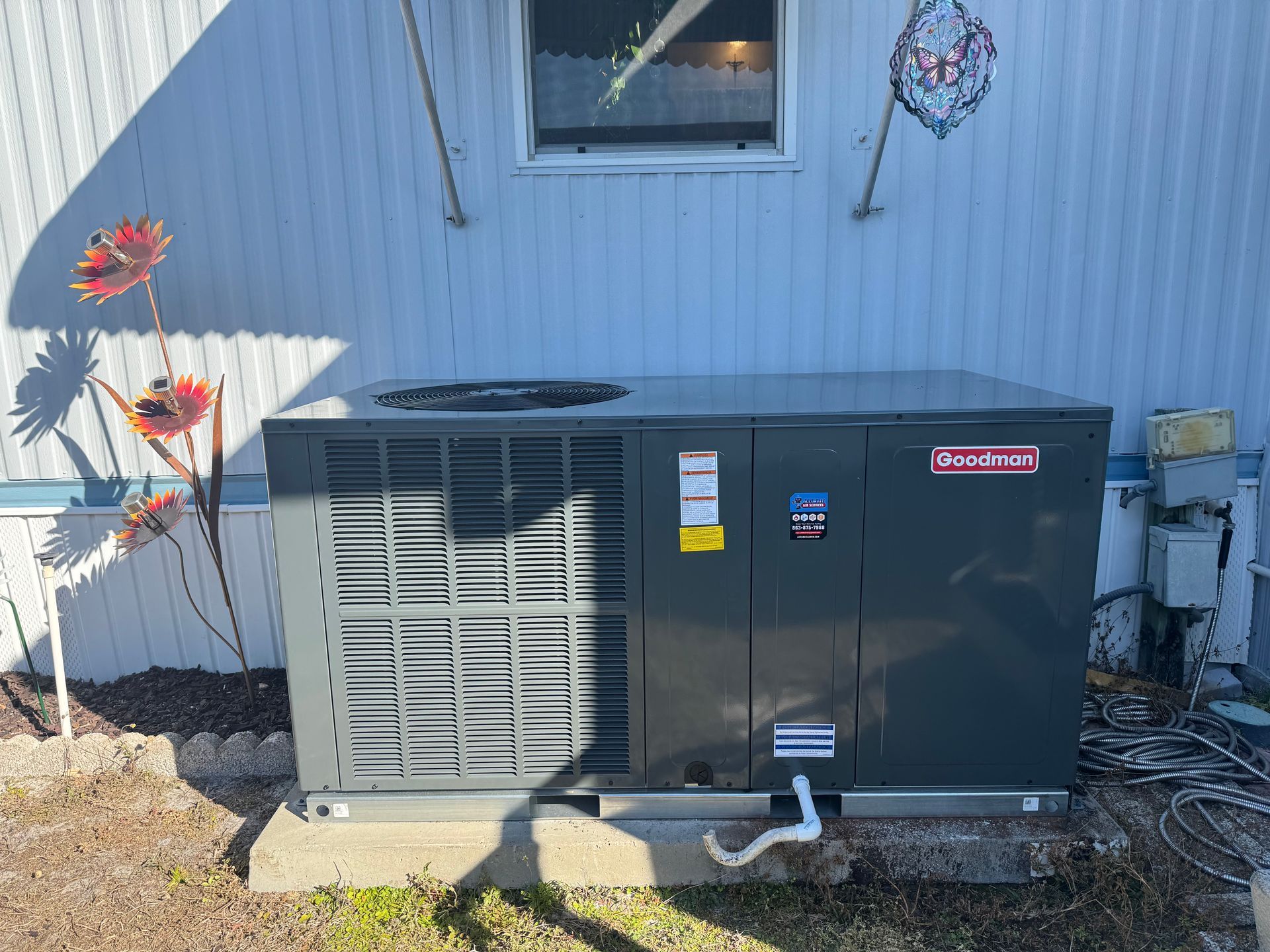 Outdoor HVAC unit beside a white house wall with decorative flowers and utility meters.