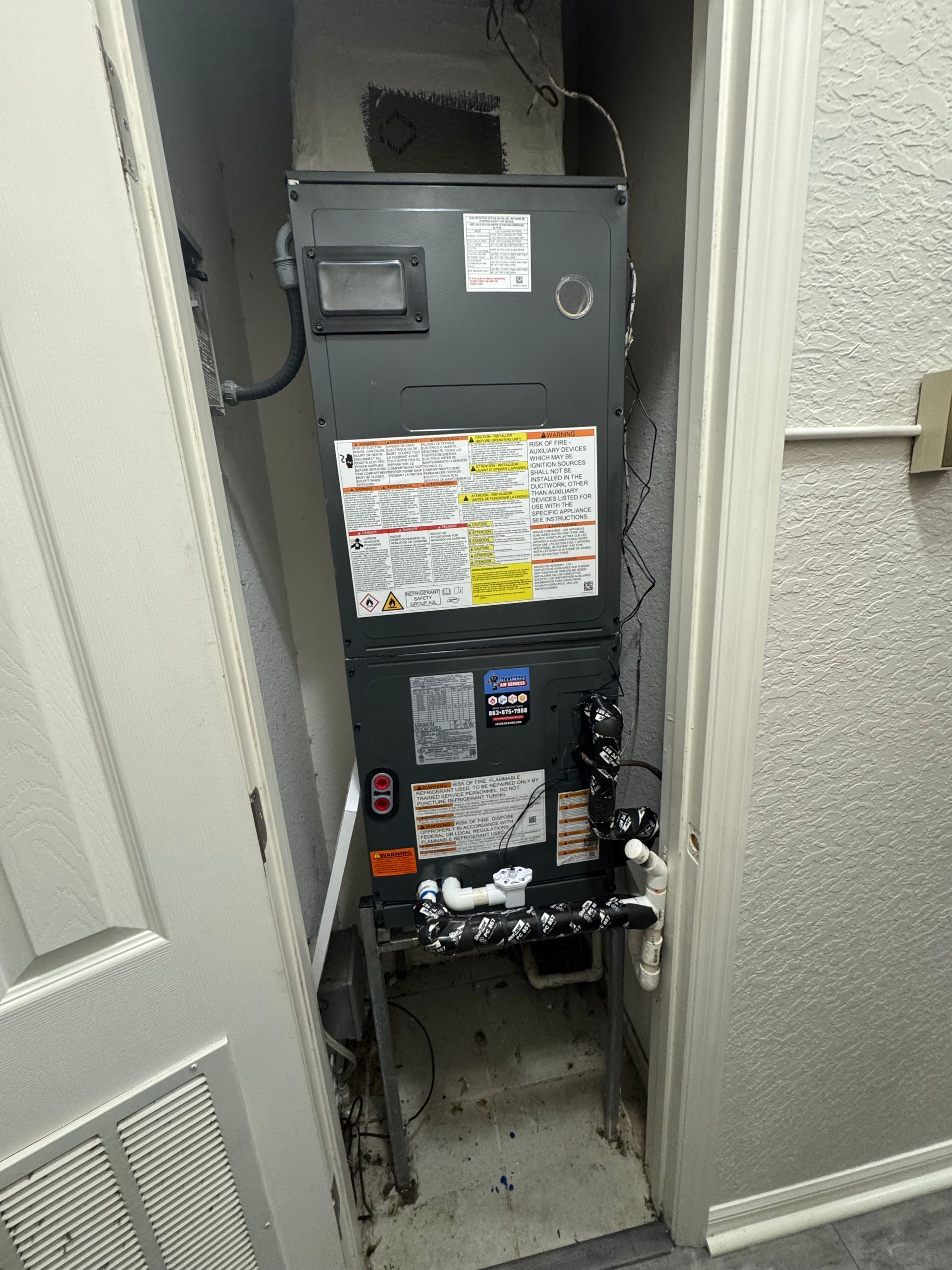 Open HVAC closet with a large black furnace or air handler and visible pipes, wires, and labels.