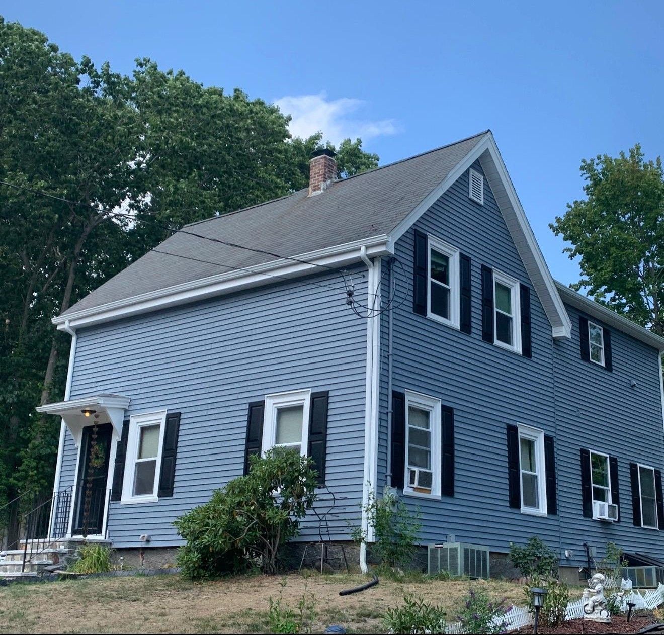 Blue-gray two-story house with white trim and black shutters, seen from the front amid trees and yard.