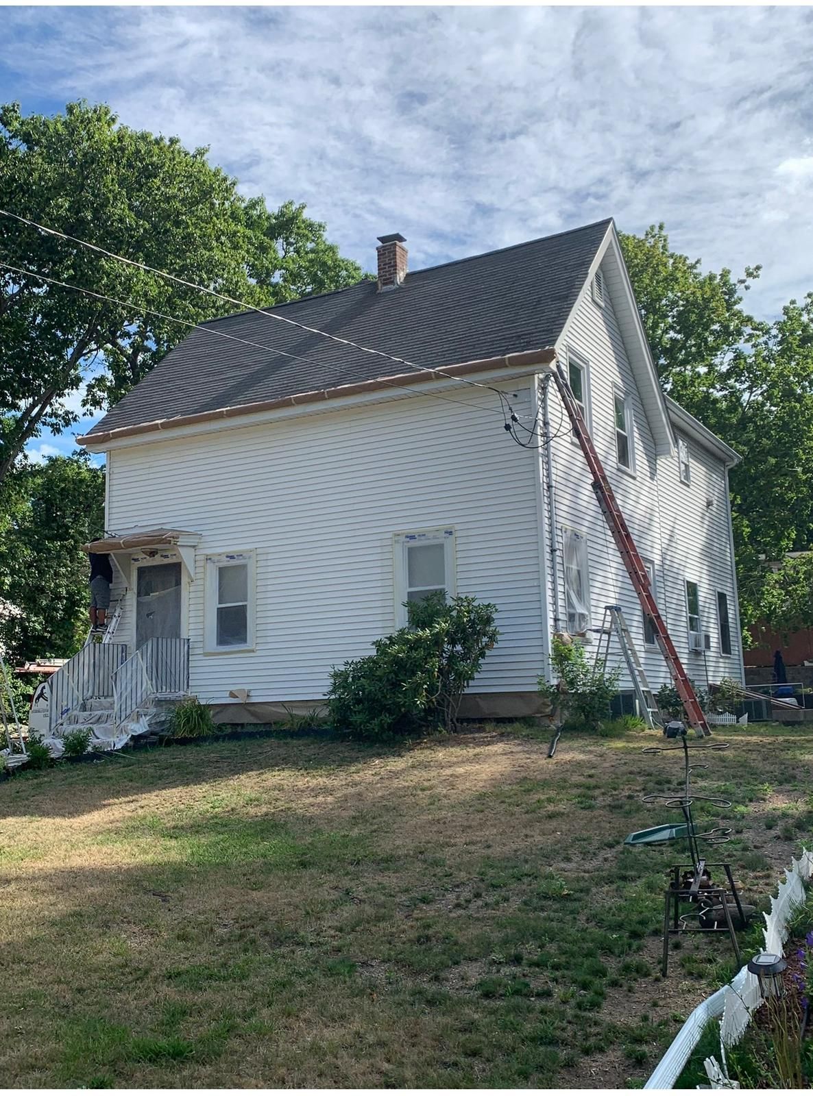White two-story house with dark roof and white porch, surrounded by trees and a grassy yard; a ladder leans on the side.