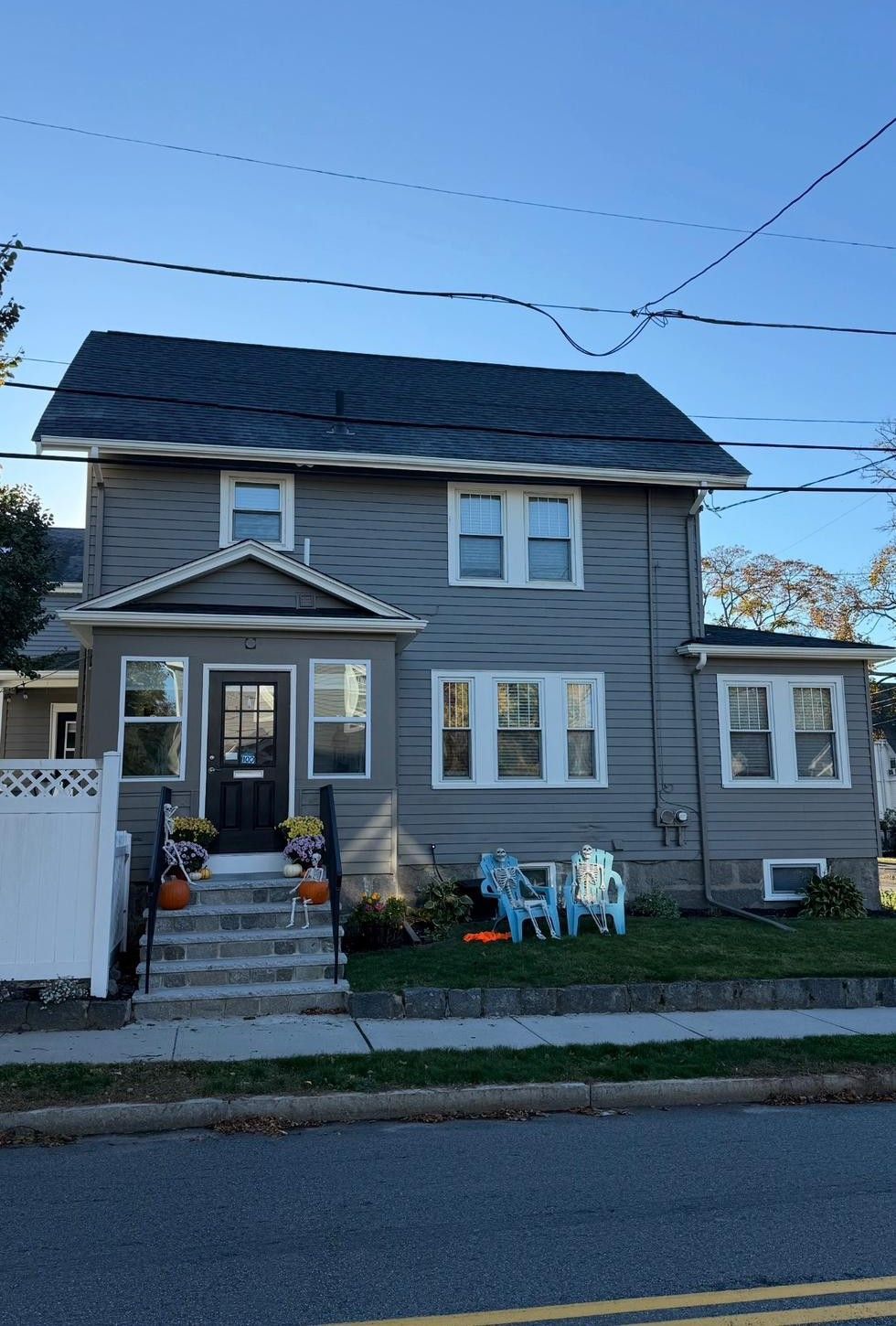 Gray two-story house with front porch, white trim, and blue lawn chairs in a neighborhood