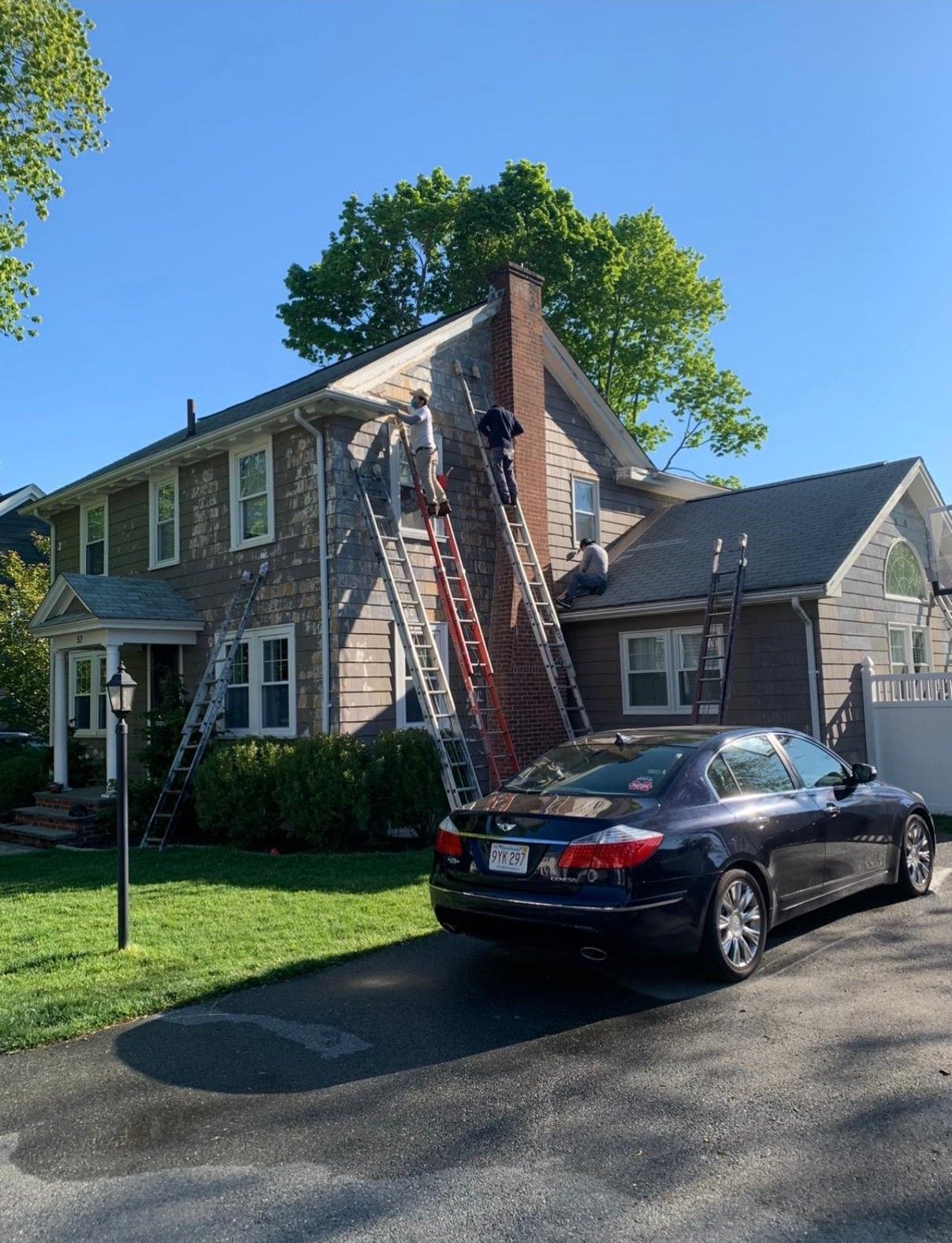 Two-story house with a tall ladder on the roof and a black car parked in the driveway under a blue sky.