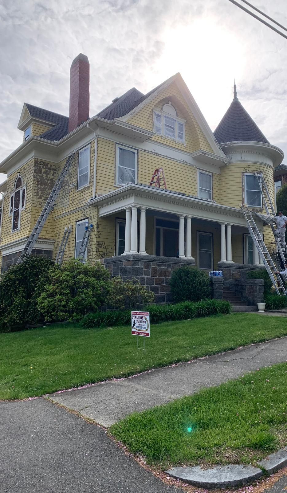 Victorian yellow house with porch, turret, and stone steps on a grassy corner lot