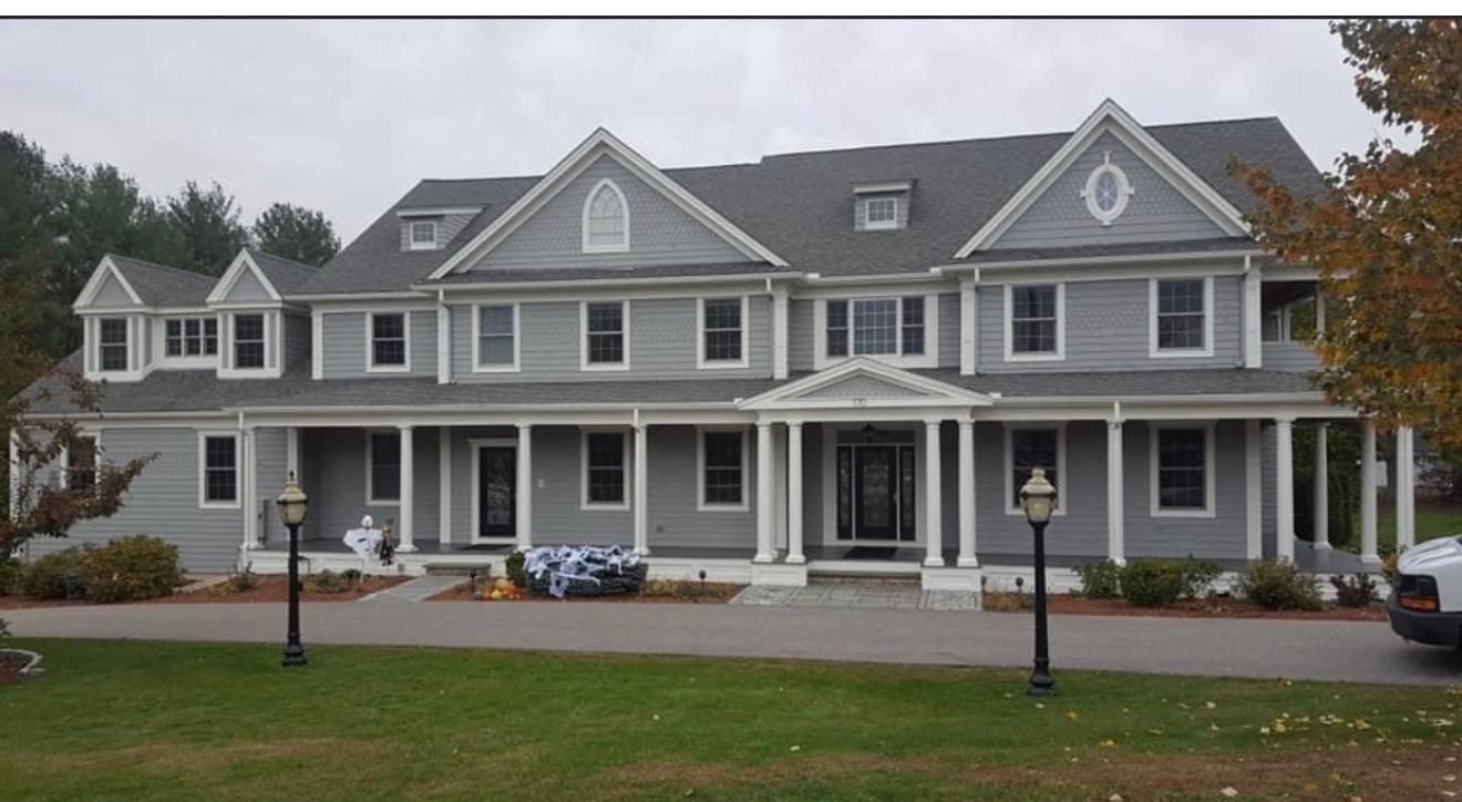 Large gray two-story house with white trim, a wraparound porch, and parked cars under an overcast sky