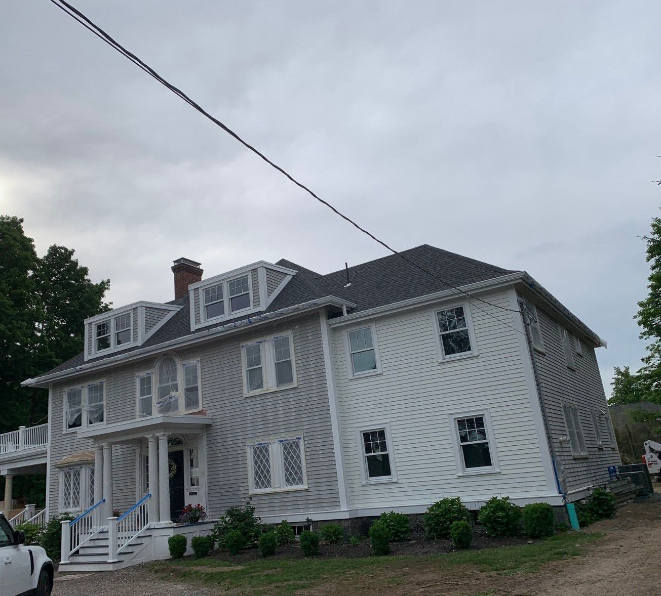 White two-story house with gray roof, front porch, dormer windows, and a gravel yard under cloudy sky