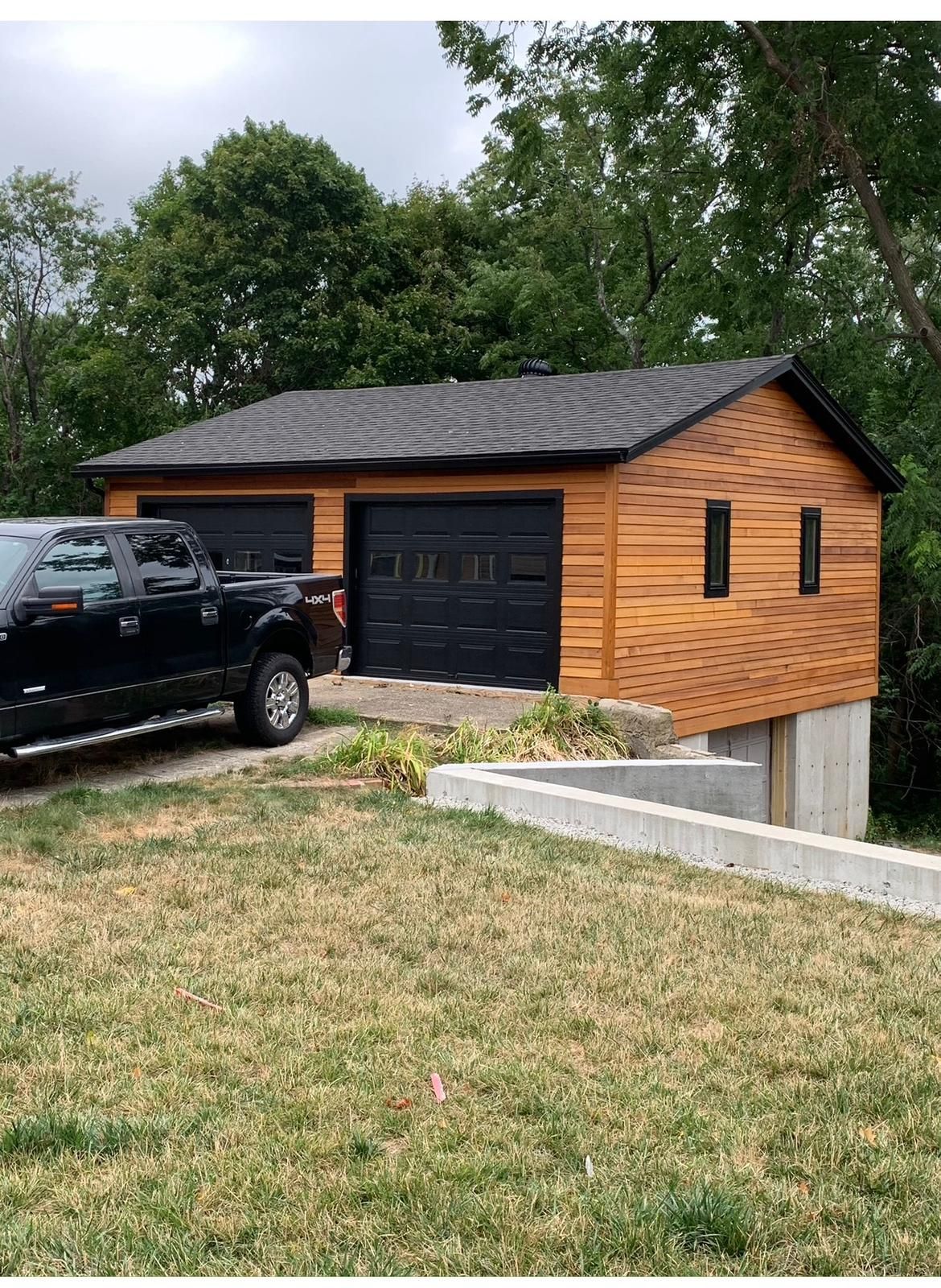 Black garage with wood siding on a sloped driveway beside a black pickup truck and trees