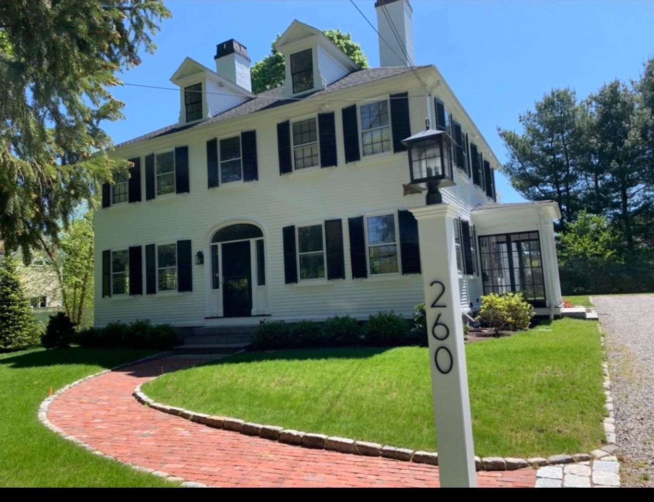 White two-story house with black shutters, red brick walkway, and address post 260 in a sunny yard
