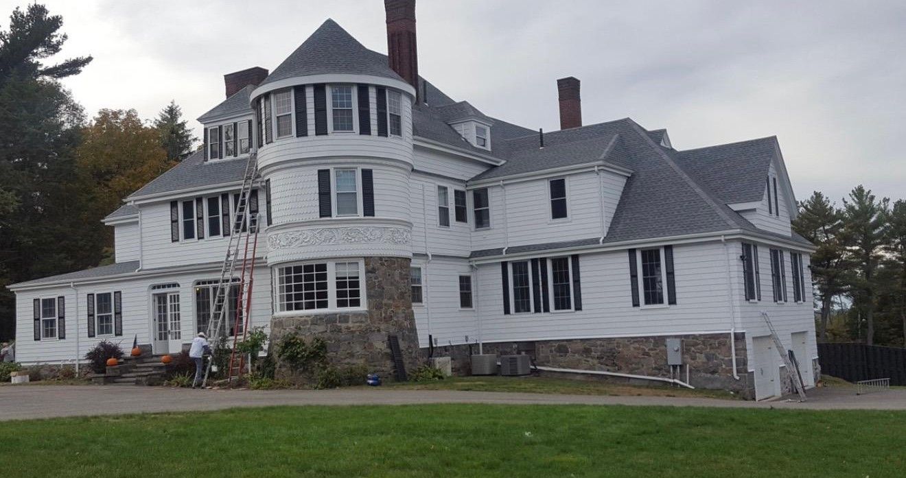 Large white Victorian house with a turret and gray roof on a grassy lawn under an overcast sky