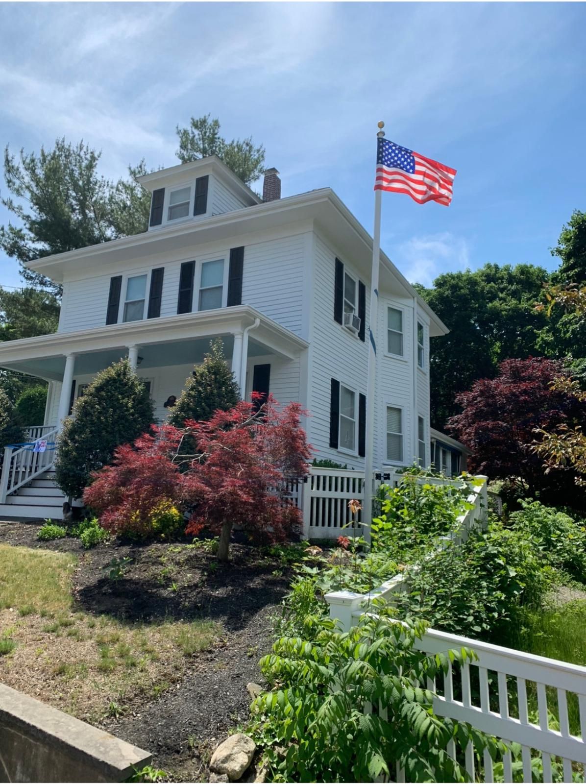 White house with American flag, surrounded by green shrubs and red flowering bushes on a sunny day.