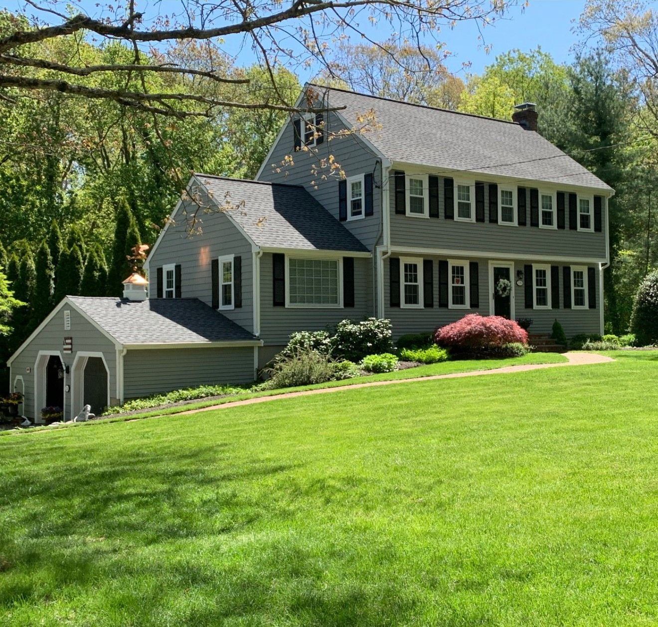 Gray two-story house with white trim on a sunny hillside, surrounded by green lawn and trees.