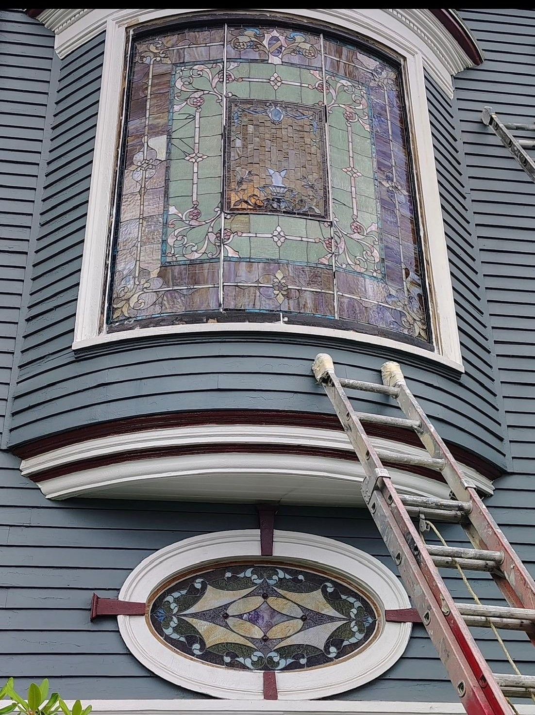 Stained glass bay window on gray house with a ladder leaning below it