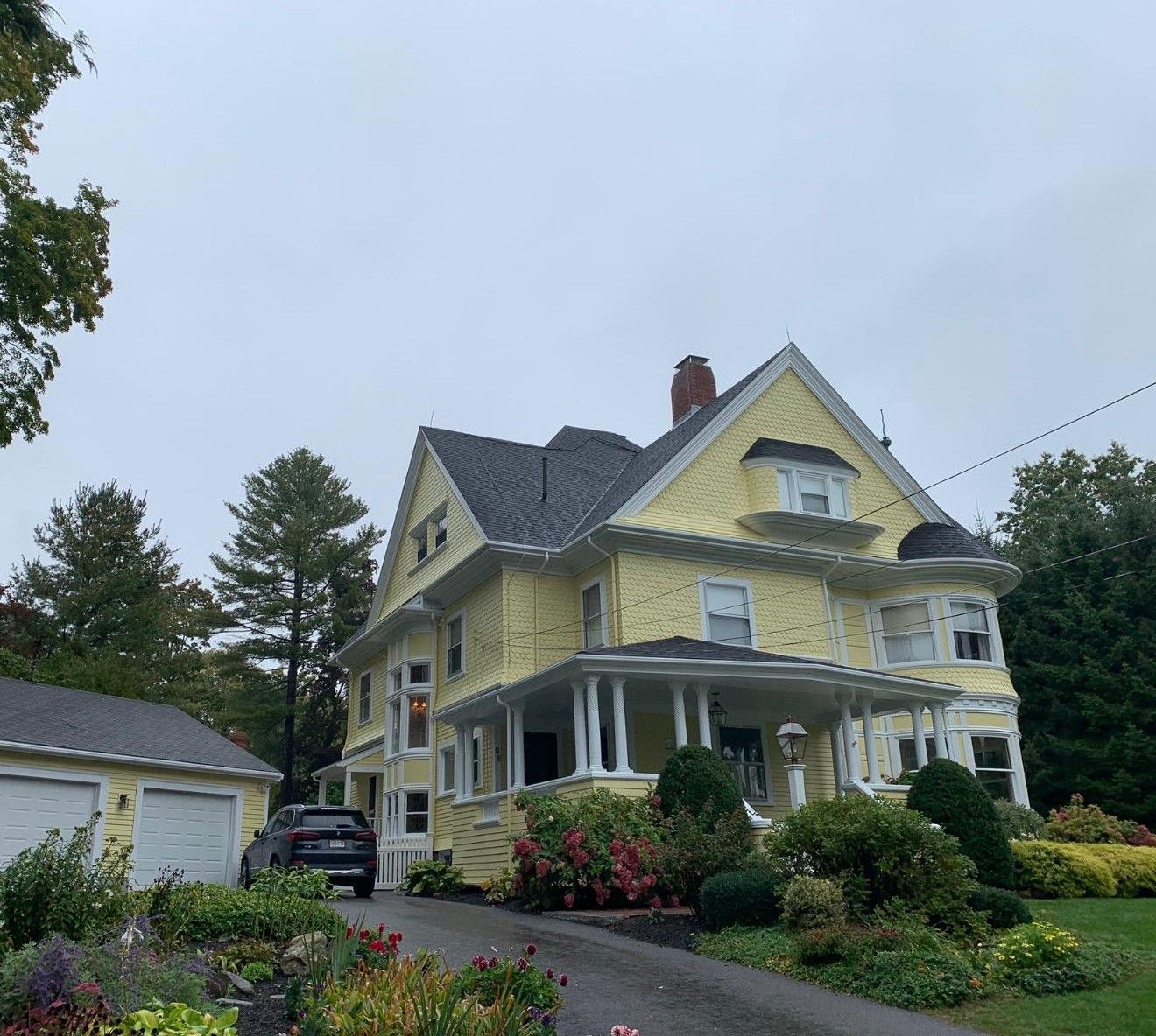 Yellow Victorian house with white trim, front porch, and lush garden beside a driveway and garage