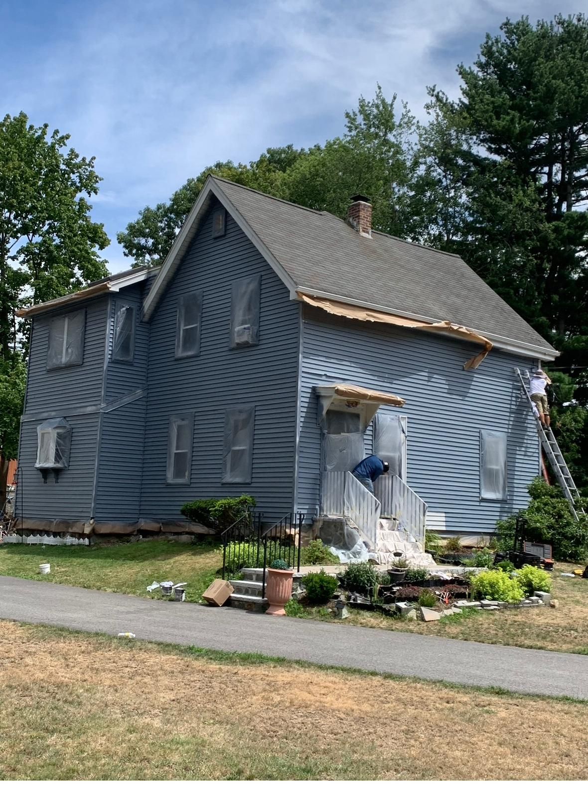 Two-story house wrapped in blue tarp with garden and lawn in front under a partly cloudy sky