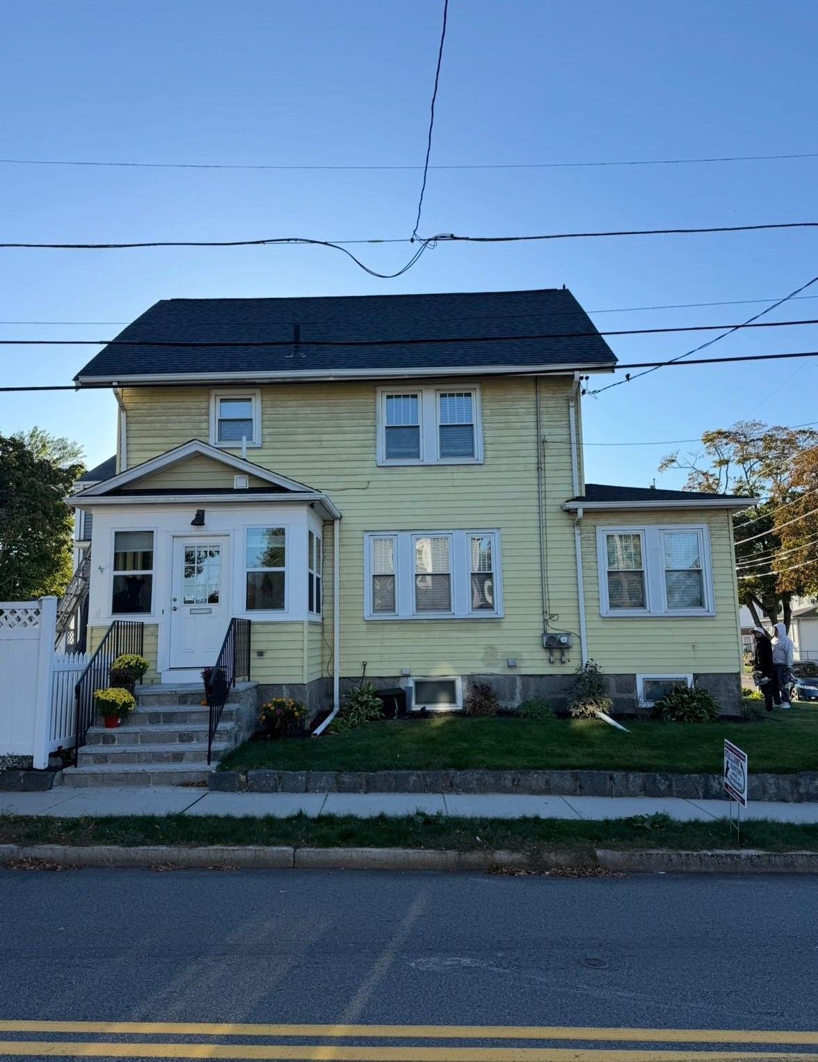 Yellow two-story house with a porch, white trim, and dark roof beside a street on a clear day