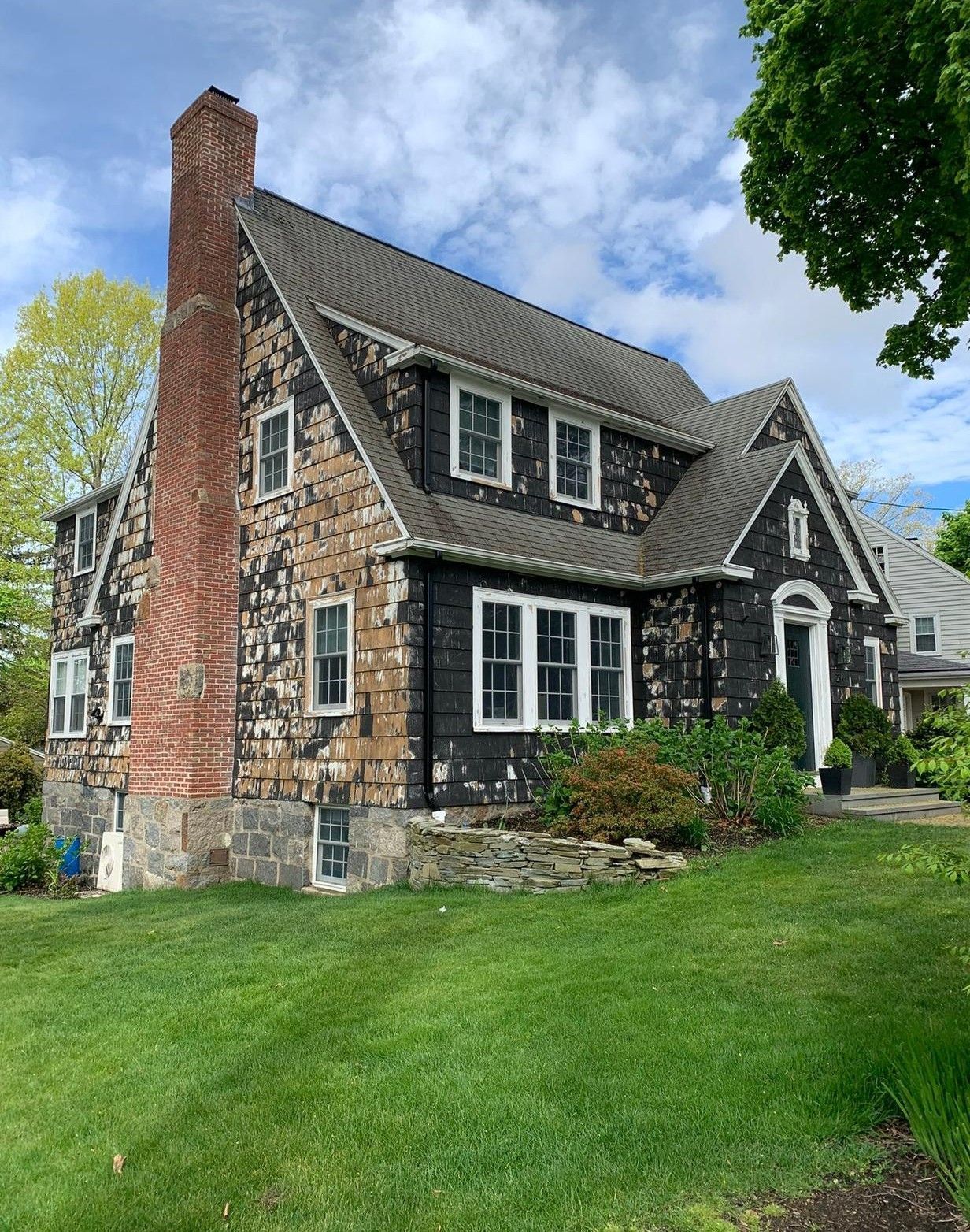 Stone-and-brick house with steep roof, tall chimney, and white-trimmed windows beside a green lawn.