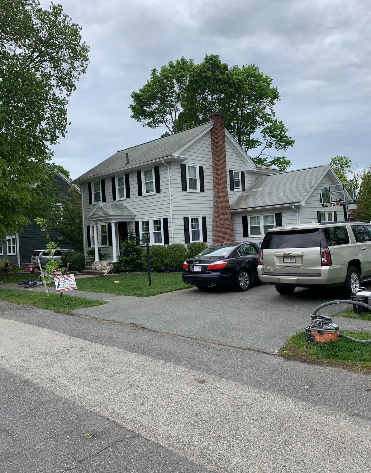 White two-story house with black shutters, parked cars, and a cloudy suburban street