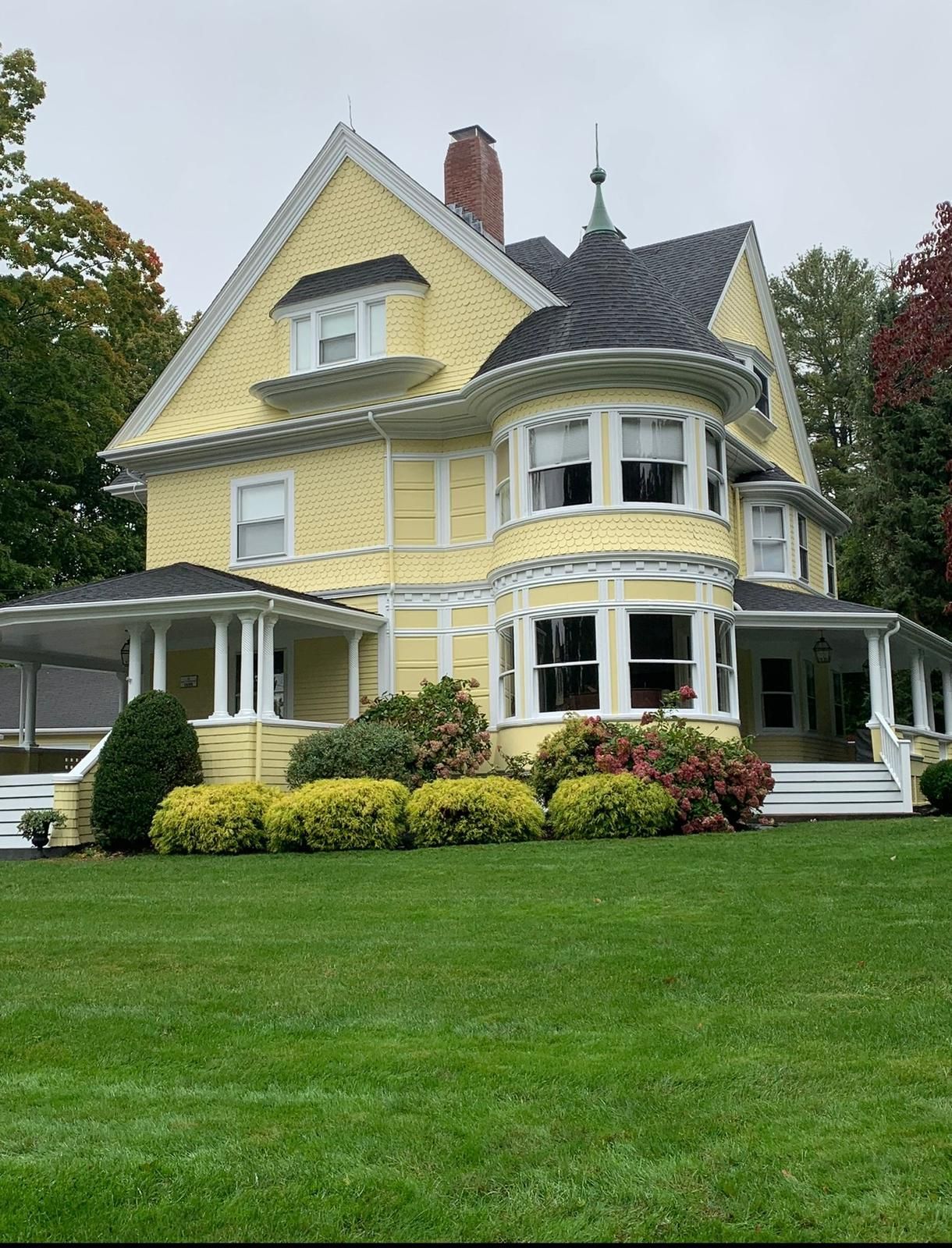 Yellow Victorian house with white trim, wraparound porch, and manicured lawn under an overcast sky