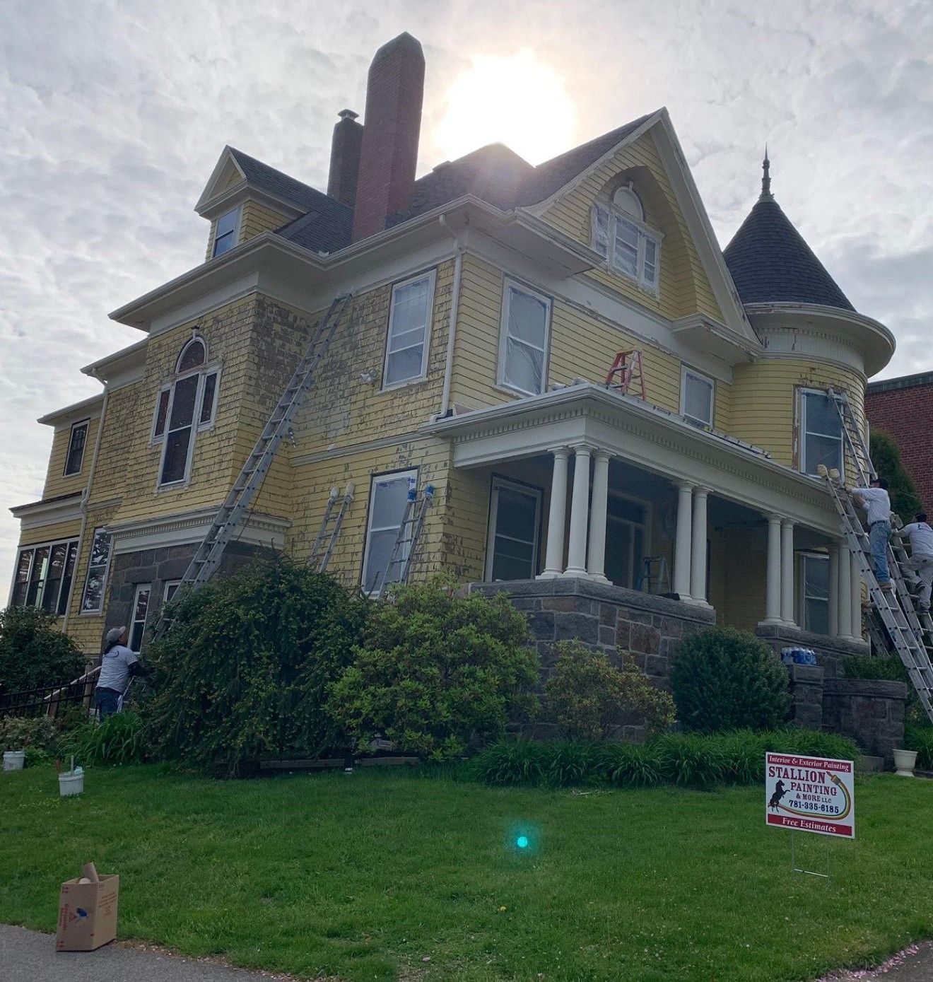 Victorian house with yellow siding, stone foundation, and a turret, viewed from a grassy front lawn.