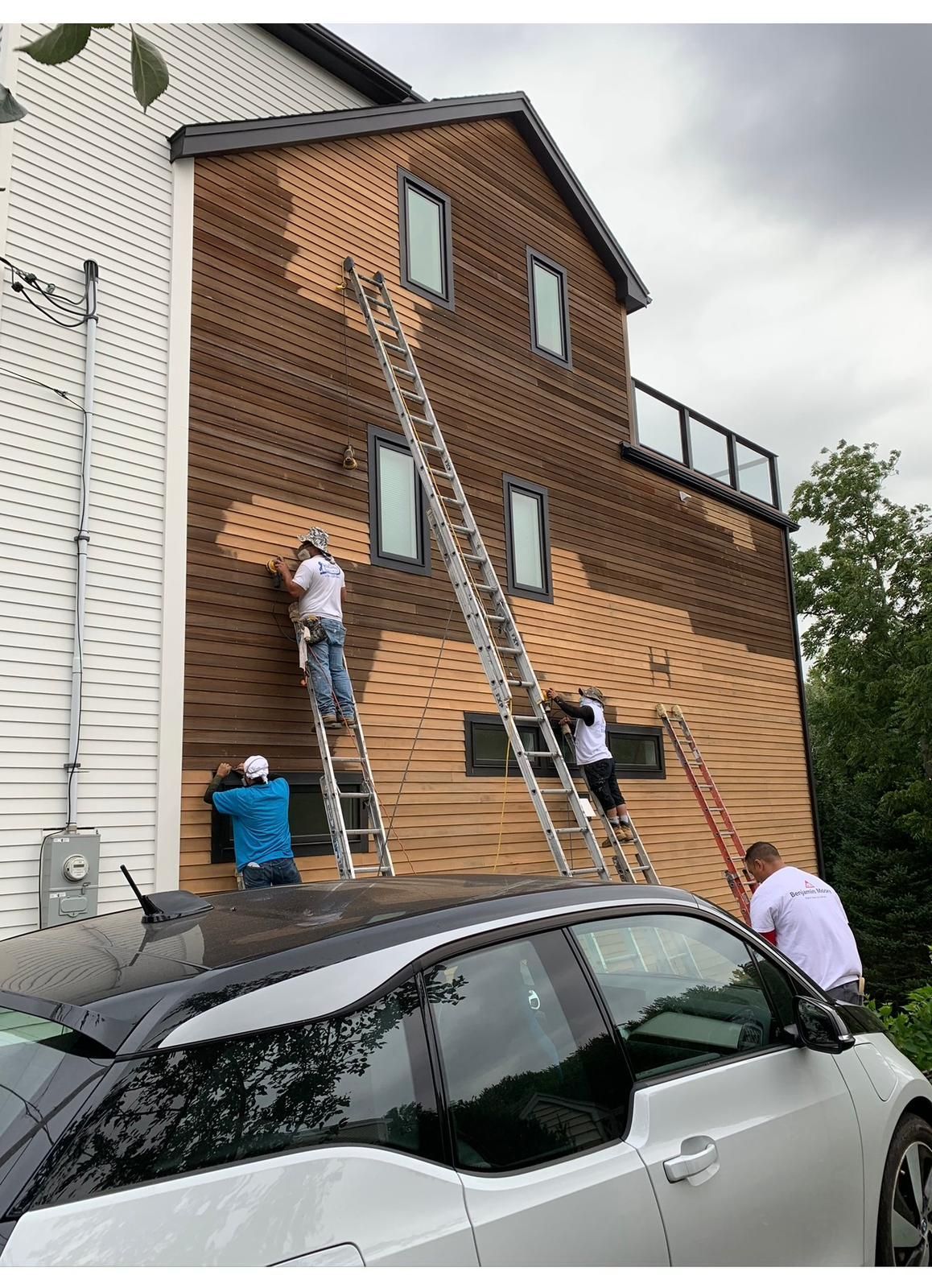 Workers on ladders painting a brick building beside parked cars