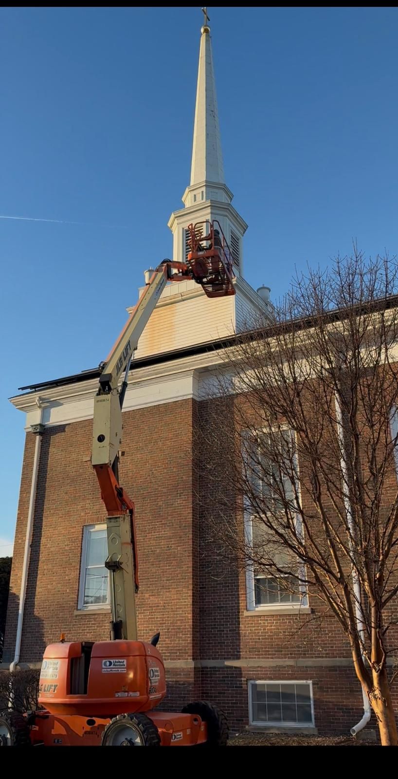 Brick church tower with a crane lifting a tall white steeple, partly framed by bare trees.