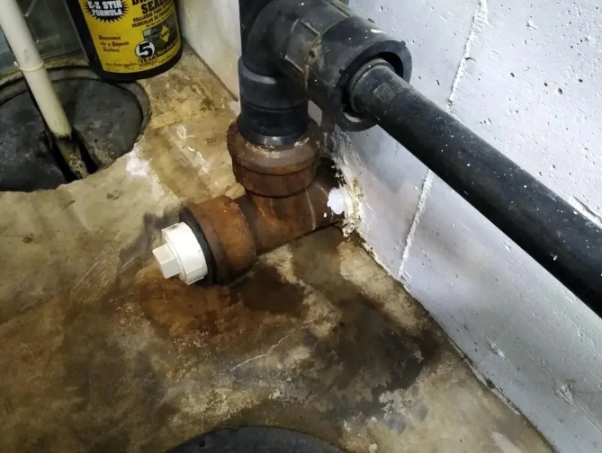 Rusty pipe connection with white fittings near a concrete wall; wet floor.
