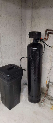 Water softener system in a concrete basement corner. Black tank, salt container, and copper pipes.