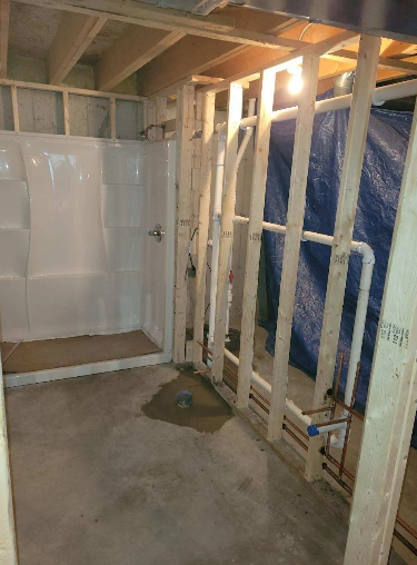 Interior of a basement bathroom under construction; shower stall, plumbing, wooden studs, concrete floor, tarp.