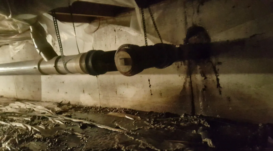 Underneath a building, a rusty pipe is connected to a larger, dark pipe. Plastic sheeting and mud are visible.