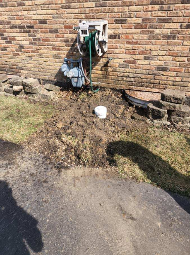 Brick wall with hose reel, exposed plumbing, and muddy ground.