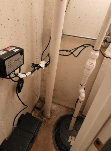 Sump pump system in a basement corner: white pipes, black pump, control box, and electrical cords against a beige wall.
