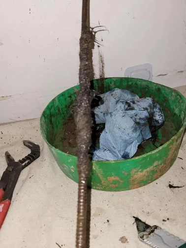 A corroded metal pipe with debris next to a green basin containing oily rags and a wrench.