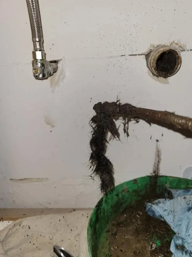 A clogged drain with a large clump of dark hair and debris being pulled out into a green bucket.