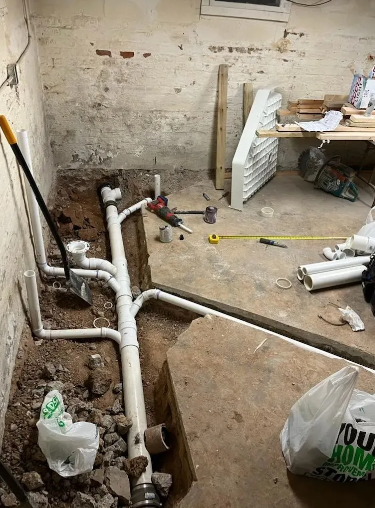 Plumbing pipes in a trench in a basement. Construction tools and supplies are scattered around the work area.
