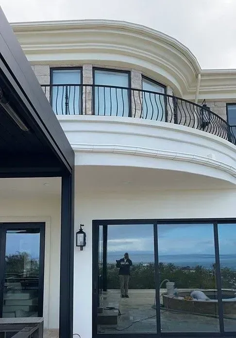 Curved white mansion with black trim and a wrought-iron balcony. A person is reflected in a large glass door, taking the photo.