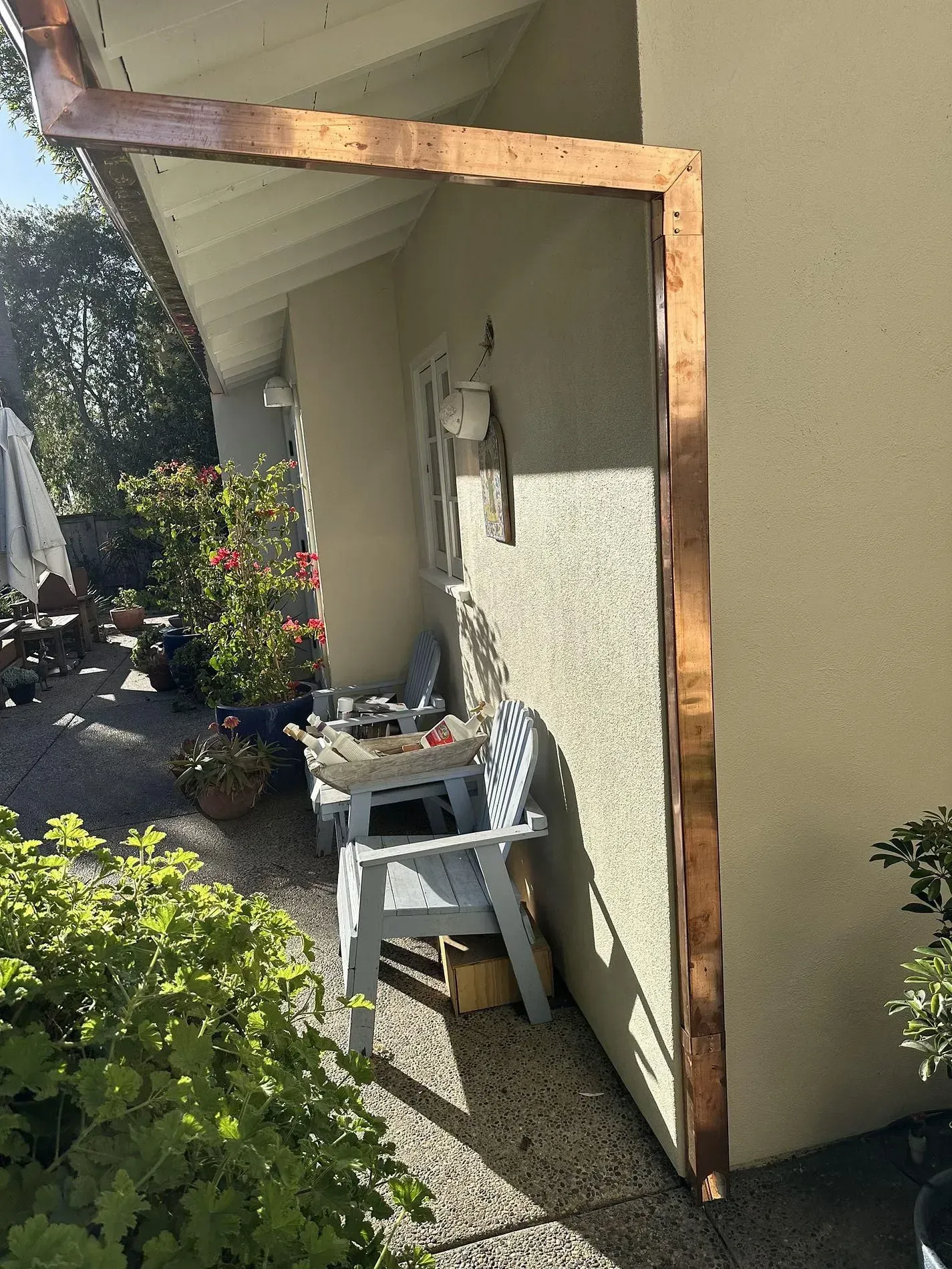 Copper flashing around a corner of a house protects the exterior. The house has a light tan stucco finish and is next to a small seating area with blue chairs.