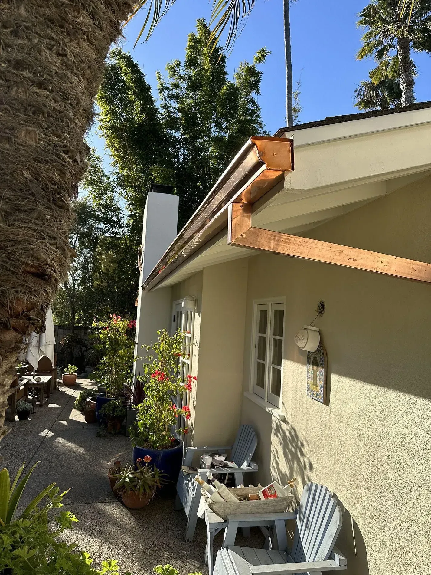 Copper gutters on a beige house, with white trim around windows. Blue chairs and plants are on a patio.