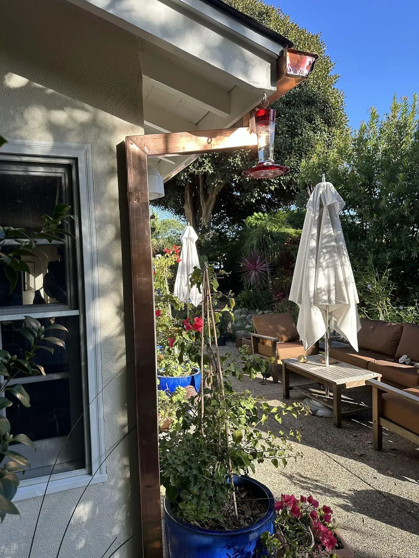 A sunny outdoor patio with a white umbrella, plants, and seating next to a house with a copper gutter.