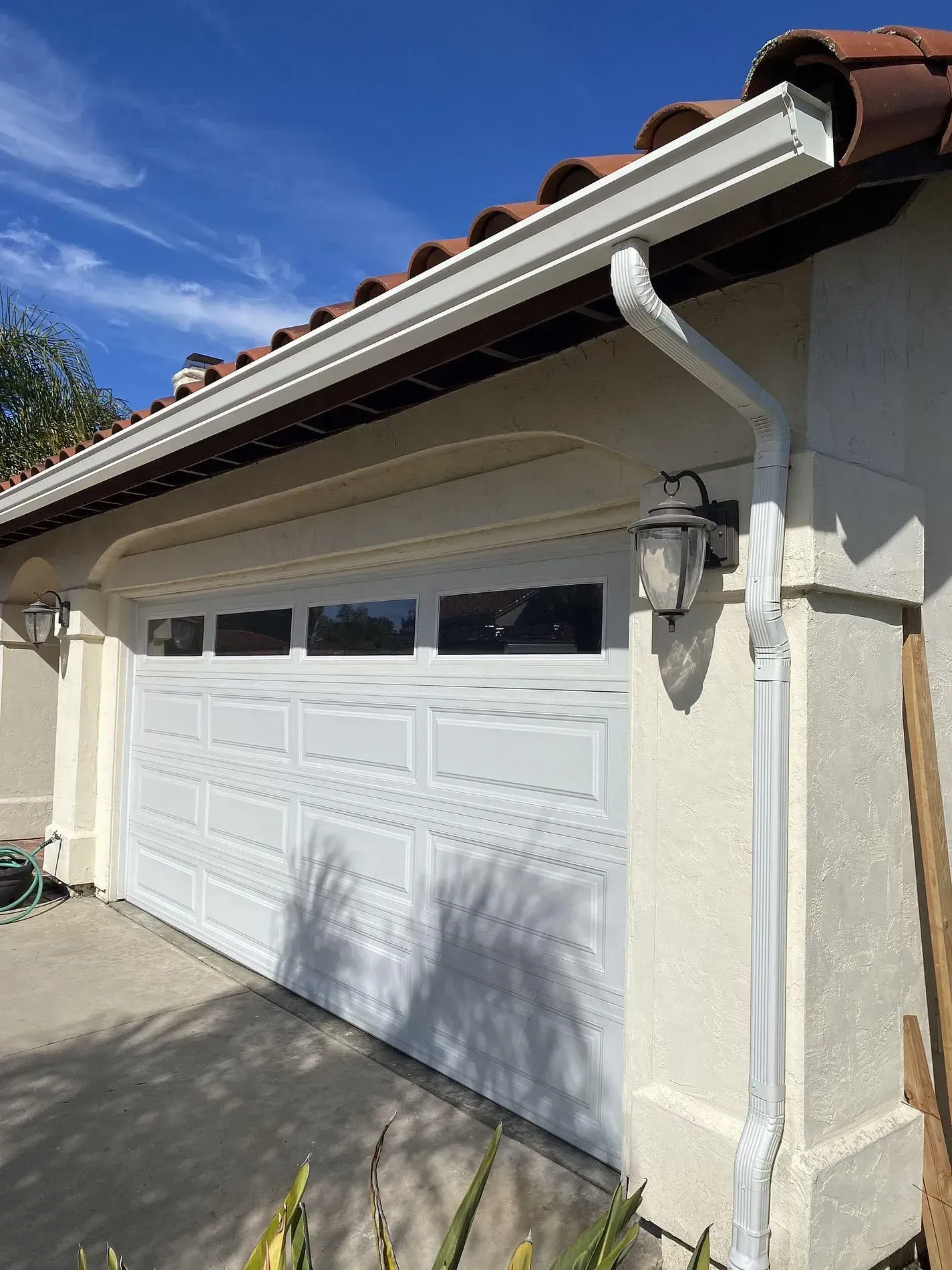 White garage door with white gutters and downspout. The stucco exterior is light-colored under a blue sky.
