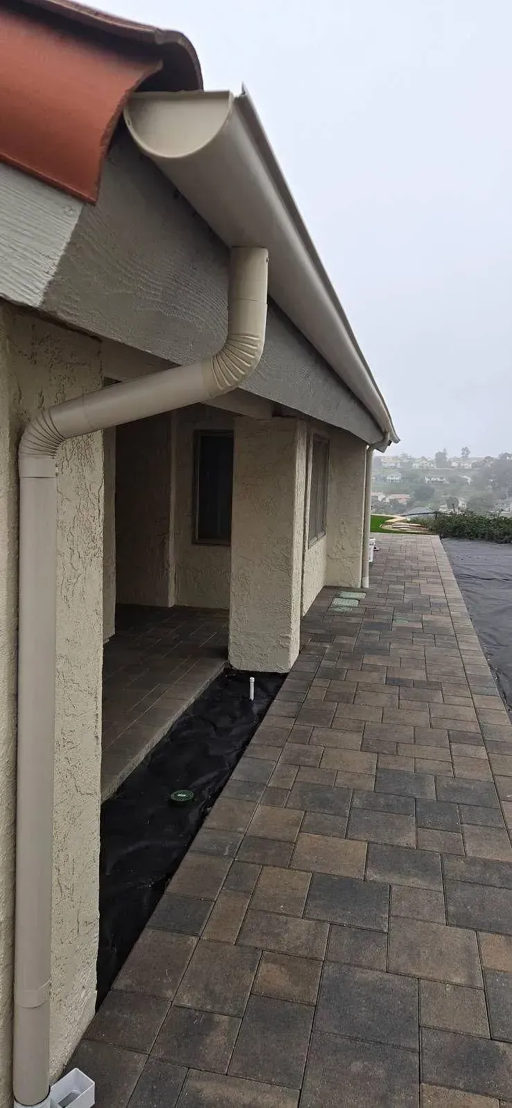 Exterior view of a building with a brown paver walkway and beige stucco walls. A gutter system is visible along the roofline.