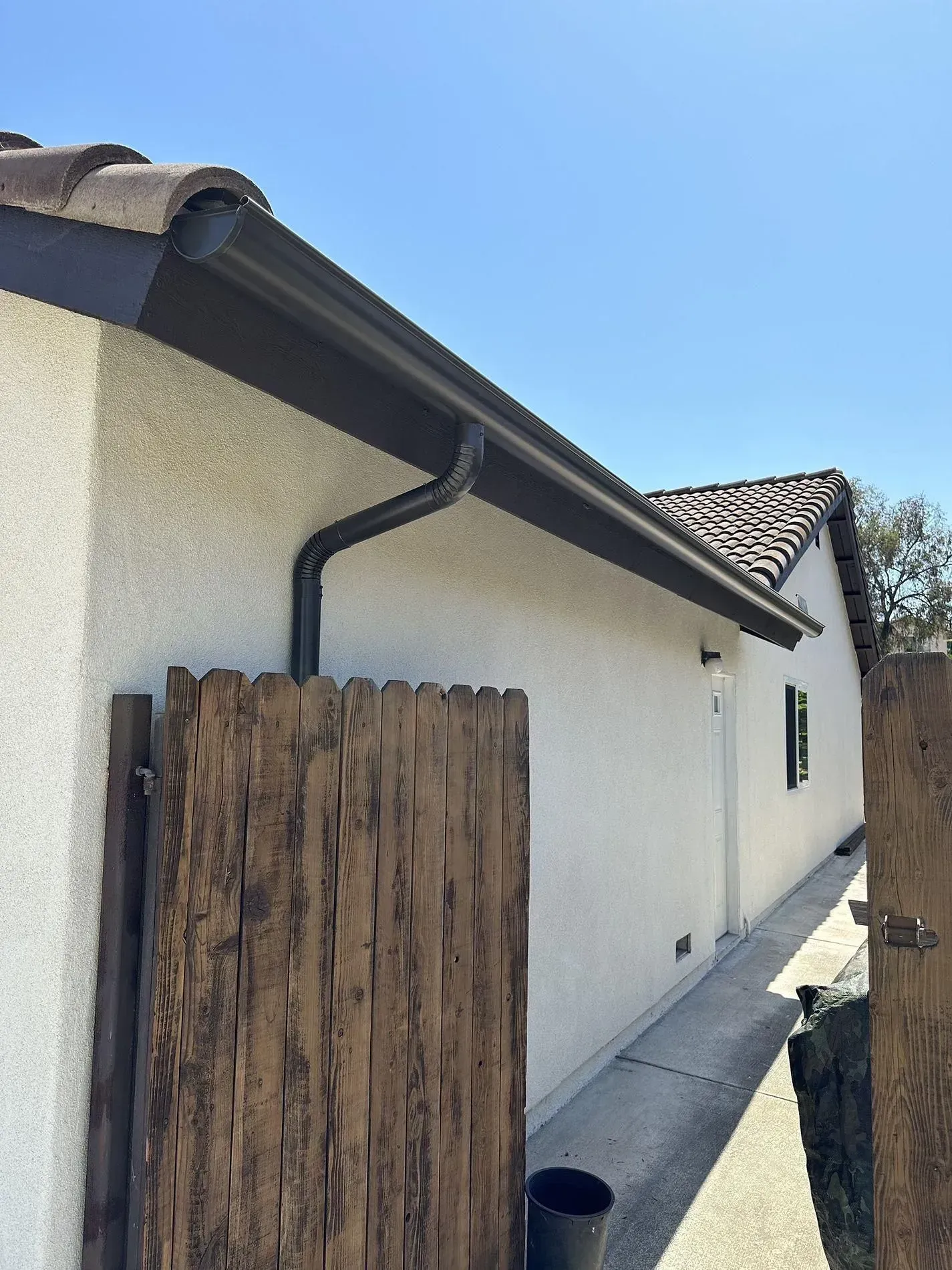 A building with a brown gutter, brown roof tiles, and a wooden fence against a clear blue sky.