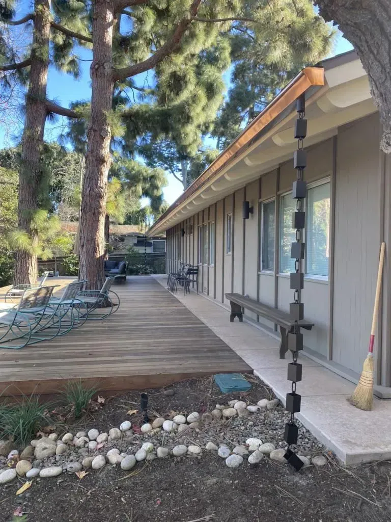 Exterior view of a building with a rain chain. Wooden deck and benches are along the side, trees in the background.