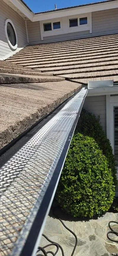 Gutter with mesh guard on a house with brown tile roof and green bush.  Black electrical cords on the ground.