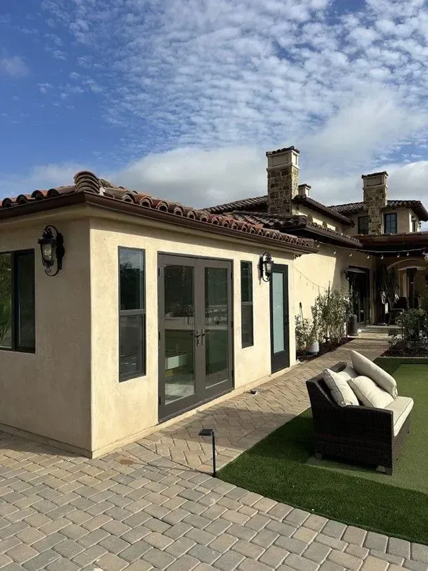 Exterior of a light-colored building with dark-framed doors and windows, a tiled roof, and a paved patio with a sofa. Green lawn and blue sky.