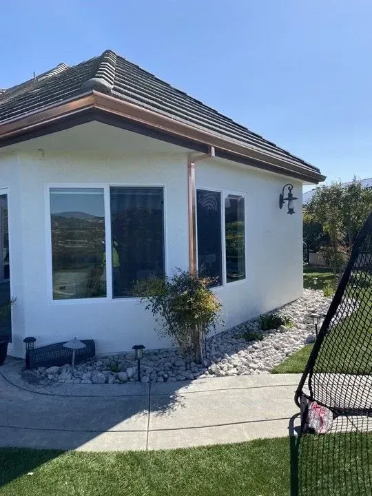 White house exterior with copper gutters, windows, and a small bush along a walkway. The sky is blue.