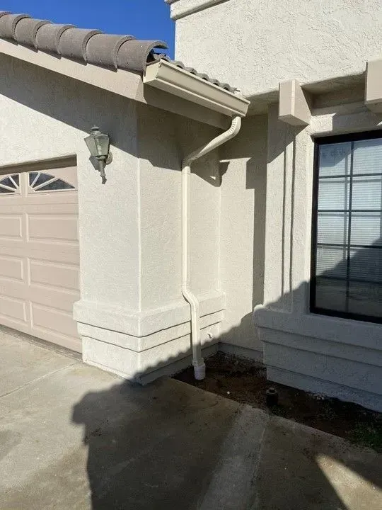 Beige house exterior with a garage door and window. A light-colored gutter runs down the wall, guiding rainwater.