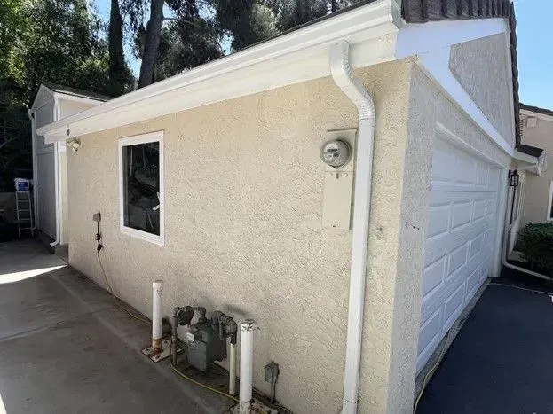 Beige stucco building with white trim and a garage door, set against a sunny backdrop. Features a window, gutters, and a gas meter.
