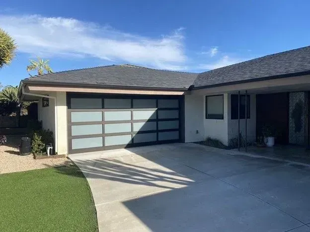 Exterior view of a house with a modern garage door. The door is glass with black frames, set in a concrete driveway, under a blue sky.