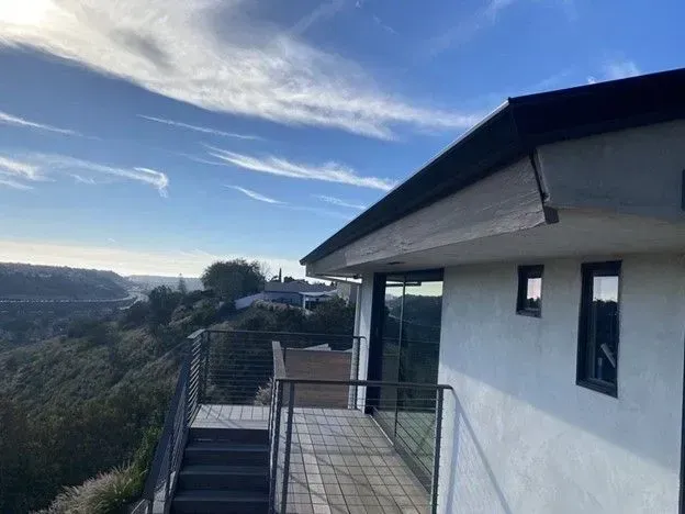 A modern house with a deck overlooking a valley, under a blue sky with clouds.