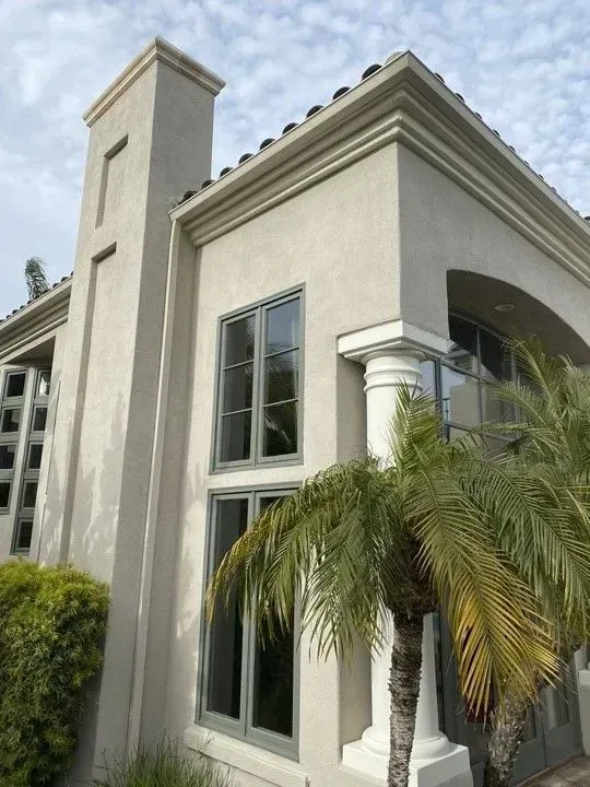 Corner of a stucco house with large windows, a palm tree, and architectural details against a cloudy sky.