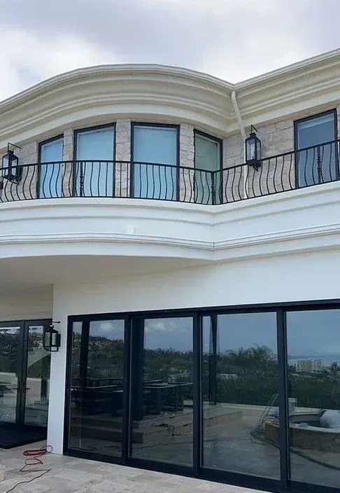 Exterior view of a modern two-story house with large glass doors and a balcony with black railings. Gray skies in the background.